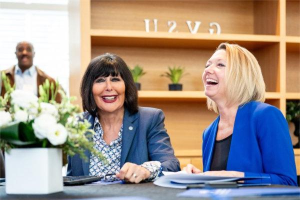 Two professionally dressed women sit at a table together laughing.