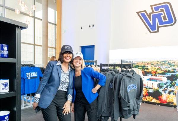 Two women in professional attire, pose together wearing baseball caps inside a store. 