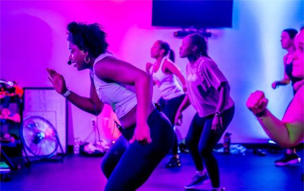 A fitness instructor leads a class in exercise in a colorful room of blue and purple lights. 