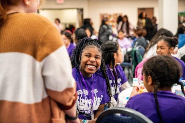 A student wearing a purple shirt smiles among a room full of event participants. 