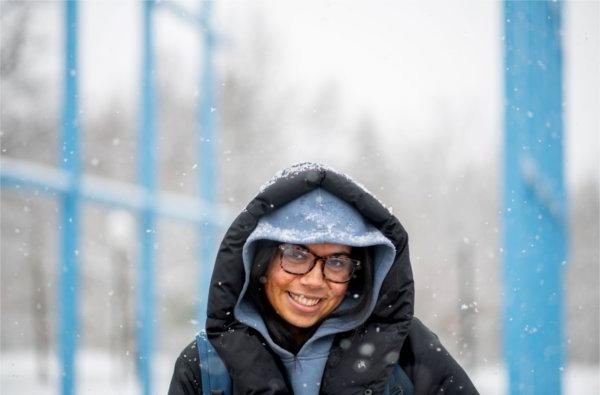 A college student smiles at the camera as snow falls on her blue and black hood and coat while walking on a college campus. 