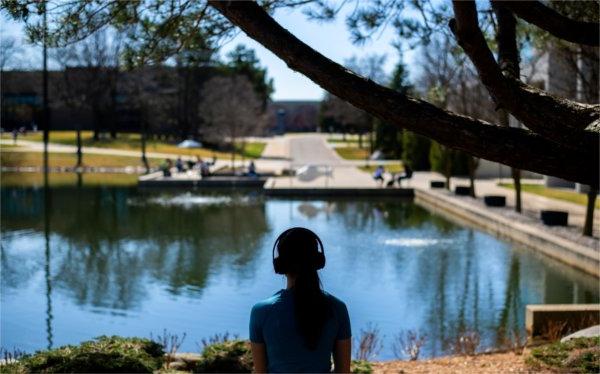 A college student is silhouetted against a pond while wearing headphones.  