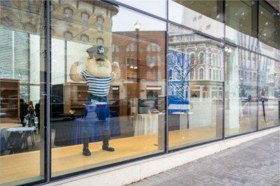 A college mascot flexes his muscles while standing in a new storefront with a city reflected in the windows.