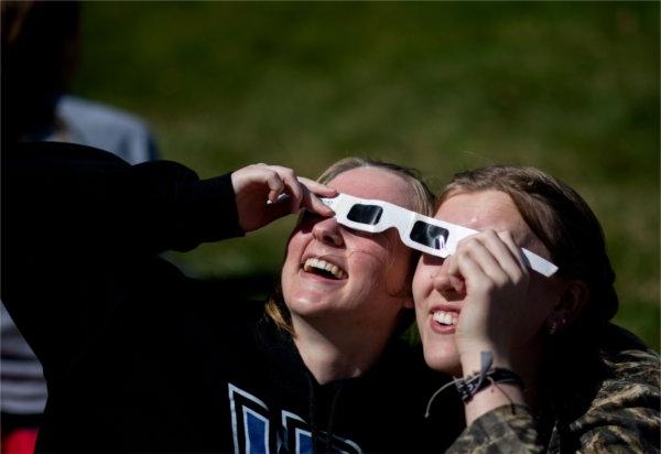 Two people smile while looking upward. They are sharing a pair of special glasses for a solar eclipse.