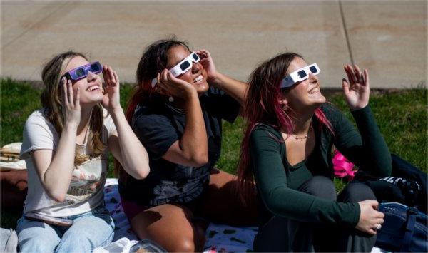 Three people sitting on the ground smile while looking upward and wearing special solar eclipse glasses.